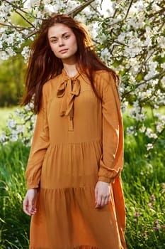 a bright vertical photo of an attractive woman in a long orange dress standing next to a flowering tree in sunny, warm weather, looking pleasantly into the camera. High quality photo