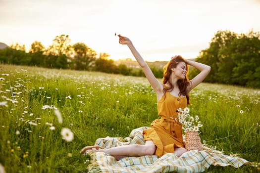 a woman in an orange dress sits on a plaid in a chamomile field and happily raises her hands up. High quality photo