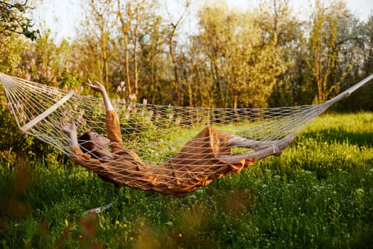 a funny woman is resting in nature lying in a mesh hammock in a long orange dress, raising her hands up, enjoying the rays of the setting sun on a warm summer day. High quality photo