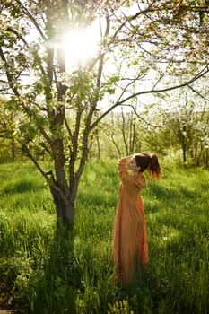 a beautiful woman stands in a long orange dress, in the countryside, near a flowering tree, during sunset, illuminated from behind and standing sideways to the camera collects her hair in a ponytail. High quality photo