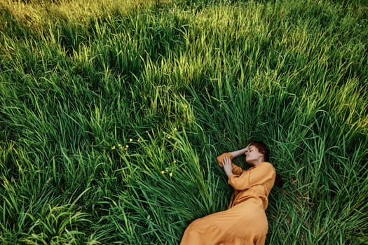 a sweet, calm woman in an orange dress lies in a green field with her hands under her head enjoying silence and peace. Horizontal photo taken from above. High quality photo