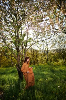 a sweet, modest, attractive woman with long red hair stands in the countryside near a flowering tree and lifts the hem of her dress up. High quality photo