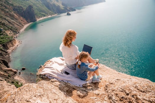 Freelance woman with her daughter working on a laptop by the sea, typing on the keyboard, enjoying the beautiful view, highlighting the idea of remote work