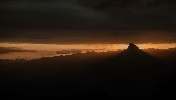 The orange horizon highlights the silhouette of the mountains as a stunning rock formation blocks the setting sun, casting rays of light over the landscape.
