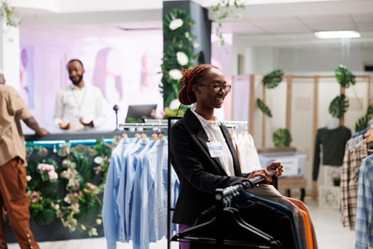 Smiling african american clothes store assistant waiting for customers while standing near apparel rack. Cheerful young fashion boutique shop professional employee working