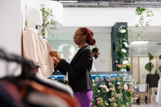 African american woman creating stylish formal look while dressing mannequin in clothing store. Fashion boutique assistant putting jacket on dummy model to showcase garment for customers