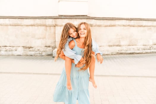 Mother of the daughter walks playing. Mother holds the girl on her back, holding her legs, and her daughter hugs her by the shoulders. Dressed in blue dresses