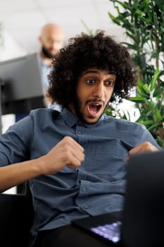 Arab company entrepreneur celebrating winning project contract while working on laptop in business office. Executive manager happy about achieving kpi, showing triumph gesture