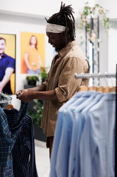 African american buyer choosing shirt and reading label on sleeve while shopping in clothing store. Man customer checking apparel before making purchase in fashion boutique