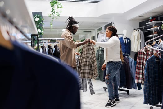 African american customer getting help from girlfriend in choosing formal shirt in fashion boutique. Couple shopping in clothing store and comparing menswear outfit on hangers