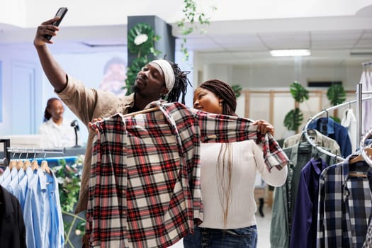 African american bloggers holding shirt and taking smartphone selfie to advertise fashion brand in store. Marketing specialist making mobile phone photo for boutique social media account