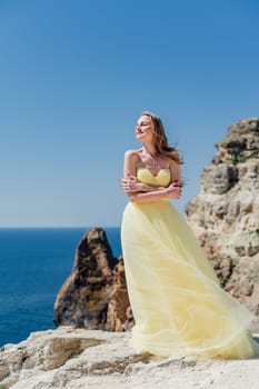 Woman in a yellow dress on the sea. Side view Young beautiful sensual woman in yellow long dress posing on a rock high above the sea at sunset. Girl in nature against the blue sky.