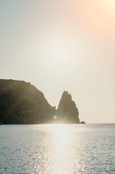 A red burning sunset with the silhouette of a cliff over the sea. Abstract nature summer or spring ocean sea background. Small waves on golden warm water surface with bokeh lights from sun.