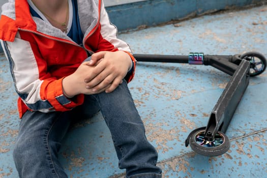 A boy holds onto a bruised knee after falling from a stunt scooter. extreme sports. do not use knee pads and elbow pads for fall protection. The concept of a healthy lifestyle and sports leisure. Child getting hurt while riding scooter. Accident with scooter