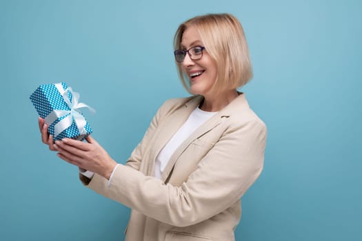 joyful 50s mature woman in jacket with gift box on studio background with copy space.