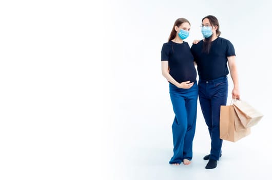 young pregnant woman and her husband with shopping bags in medical masks. safety from colds flu and coronavirus on white background. Pregnancy shopping concept happy young family with shopping bags