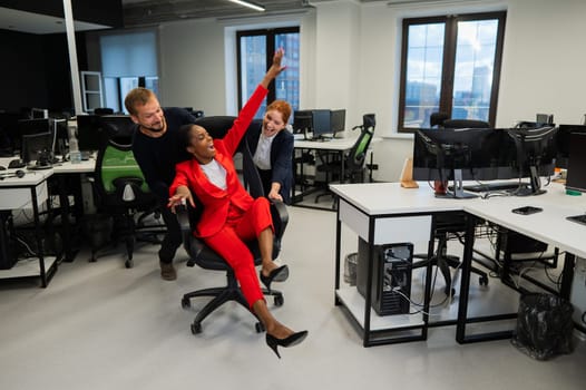 Caucasian red-haired woman, bearded caucasian man rolled African American young woman on office chair. Colleagues have fun at work