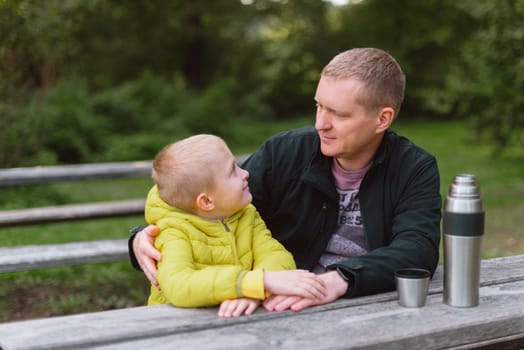 Happy family: father and child boy son playing and laughing in autumn park, sitting on wooden bench. Father and little kid having fun outdoors, playing together. Father and son sitting on a bench and talking. dad son park bench table autumn thermos.