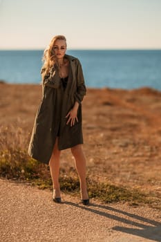 Portrait blonde sea cape. A calm young blonde in an unbuttoned khaki raincoat walks along the seashore, under a raincoat a black skirt and top.