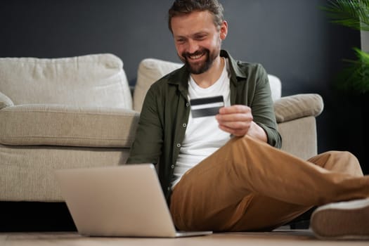 Happy, smiling customer sitting on floor next to sofa while shopping online using credit card for online auction. Concept of online payment systems, e-commerce, and online shopping in modern and technological setting. High quality photo