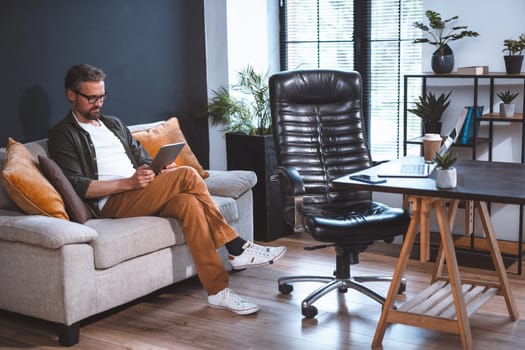 Freelancer sitting on sofa, looking relaxed as he uses tablet PC. Laptop on desk and empty chair can be seen nearby in room. Concept of relaxation after work, and ease and comfort of working from home. High quality photo