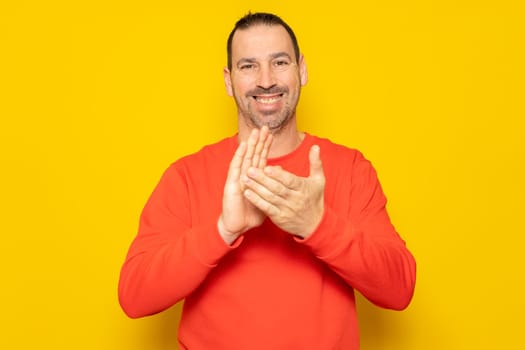 Bearded hispanic man in his 40s wearing a red jumper clapping hands happy and cheerful, smiling proud hands together. Isolated on yellow studio background