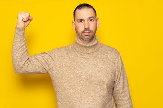 Bearded Hispanic man in his 40s wearing a beige turtleneck showing off his biceps exposing his sorry state of shape, isolated on yellow studio background