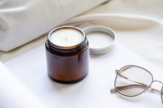 Soy wax aroma candle in brown jar on bed , with fashion glasses. Candle mockup design. Mockup soy wax candle in natural style.