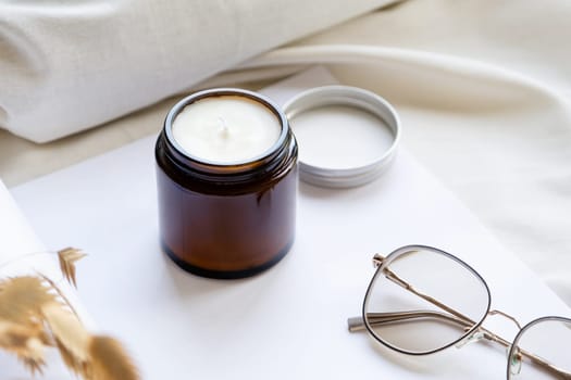 Soy wax aroma candle in brown jar on bed , with fashion glasses. Candle mockup design. Mockup soy wax candle in natural style.