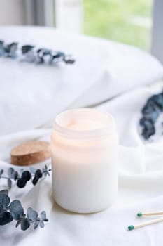Soy wax aroma candle in white jar on bed with eucalyptus leaves. Candle mockup design. Mockup soy wax candle in natural style.