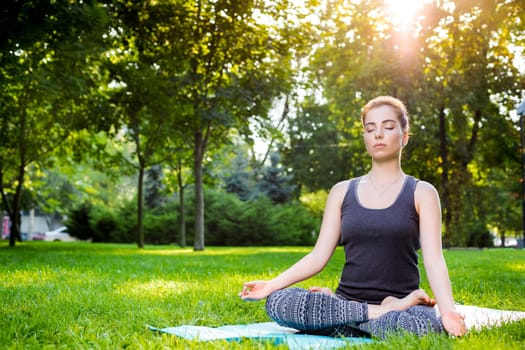 Meditating woman in meditation in city park in yoga pose. Girl relaxing with serene relaxed expression outside in summer. Beautiful young Caucasian female model. sun flare