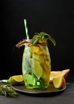 Lemonade in a transparent glass with lemon, lime, rosemary sprigs and mint leaves on a black background