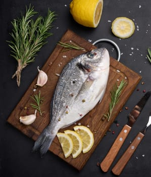 Raw whole dorado fish on wooden kitchen board and spices for cooking, top view on black table