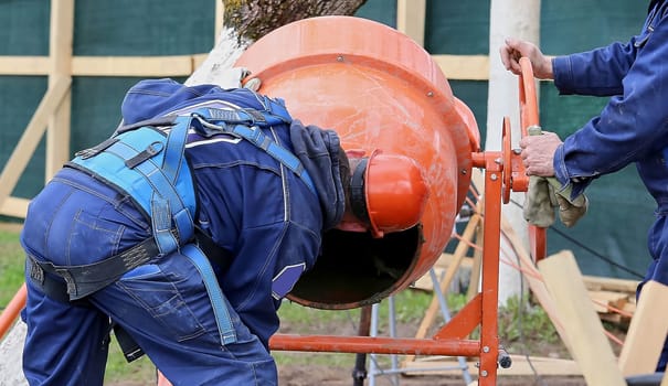 Construction of a brick house. A man in an orange construction helmet looks into a mobile concrete mixer. Concrete works