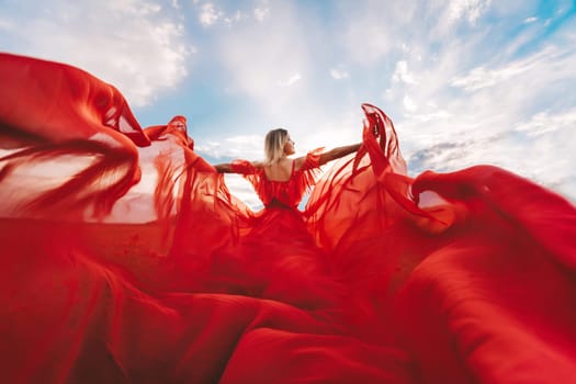 Woman Red Flying Dress. A blonde in a red dress against the sky. Rear view of a beautiful blonde woman in a red dress fluttering in the wind against a blue sky and clouds