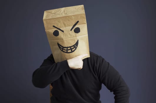 A man in a black turtleneck with a paper bag with an angry emoticon on his head shows a threatening gesture. Emotions and anger