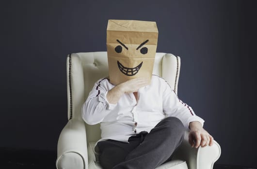 A man with a paper bag on his head, with an evil smiley face drawn, sits on a white chair, gloats emotionally. Emotions and gestures.