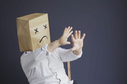 A sad man in a white shirt with a bag on his head, with a sad emoticon drawn, is afraid, defends himself by putting his hands forward. Emotions and gestures.