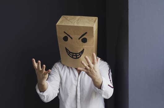 A man in a white shirt with a paper bag with an angry emoticon on his head gesticulates aggressively with his hands. Emotions and anger