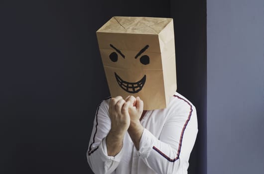 A man in a white shirt with a paper bag with an angry emoticon on his head shows a threatening gesture. Emotions and anger