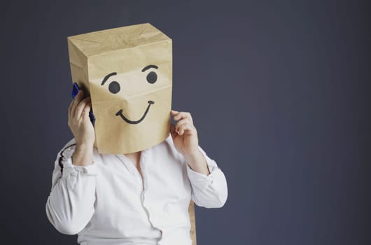 A man in a white shirt with a paper bag on his head, with a smiley face drawn, talking on the phone.