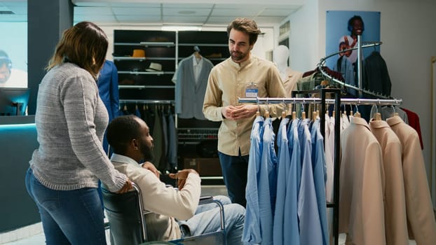 African american man with disability visiting retail shop, shopping for trendy fashion collection in clothing store. Male wheelchair user buying casual wear from market shop boutique.