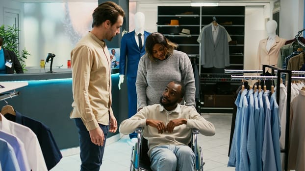 Male wheelchair user shopping for trendy clothes in fashion boutique store, looking at merchandise on hangers. Person with chronic impairment buying modern formal wear, small business.