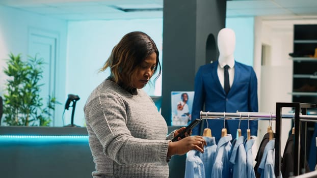 African american woman looking at website stock, trying to find clothes from online app in clothing store. Young client using smartphone to buy favorite trendy merchandise in shopping mall.