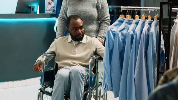 African american client in wheelchair shopping for clothes, buying formal or casual wear from trendy fashion boutique. Male person with chronic disability looking at shop merchandise.