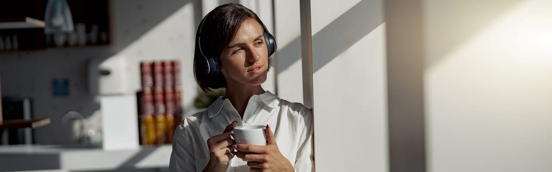 European business woman in headphones holding cup of coffee while standing near window at office