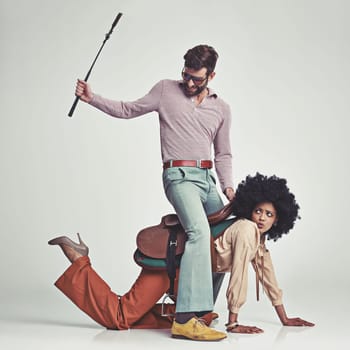 Hes in charge now. A studio shot of an attractive man in 70s wear riding a young woman wearing a saddle while using a riding crop