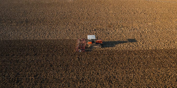 farmer driving a crawler tractor drags a cloud of dust behind, ploughing, power harrowing surface crust breaking to facilitate the birth and seeding of plants on dry soil