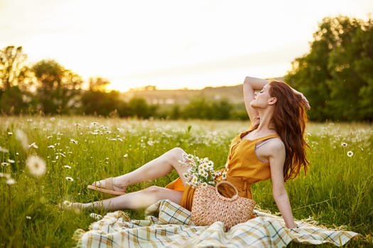 a beautiful woman in an orange dress sits on a plaid in a chamomile field and straightens her hair. High quality photo