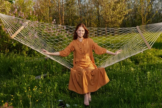 horizontal photo of a beautiful, red-haired woman lying in a hammock enjoying a rest in a long orange dress, on a warm summer day, smiling happily looking at the camera. High quality photo